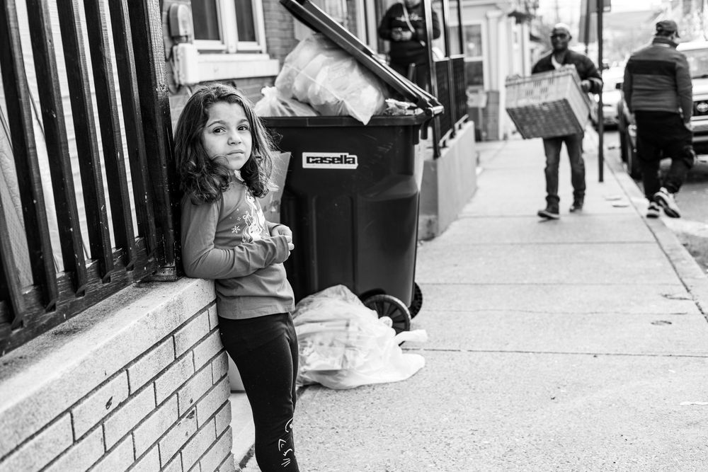 Black and white photo of a young girl leaning against a brick wall on a city sidewalk near a trash can.
