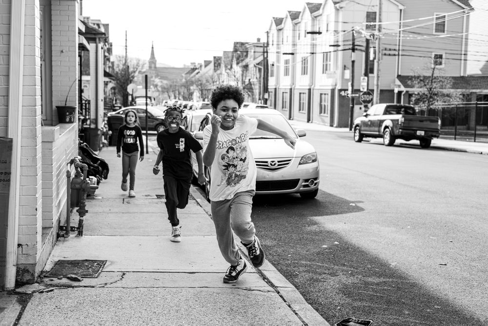 Children running and playing on a city sidewalk in a black and white urban street photography scene.
