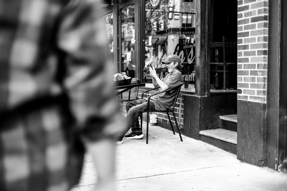 Black and white street photo of a person sitting outside a brick building cafe, looking at their phone.