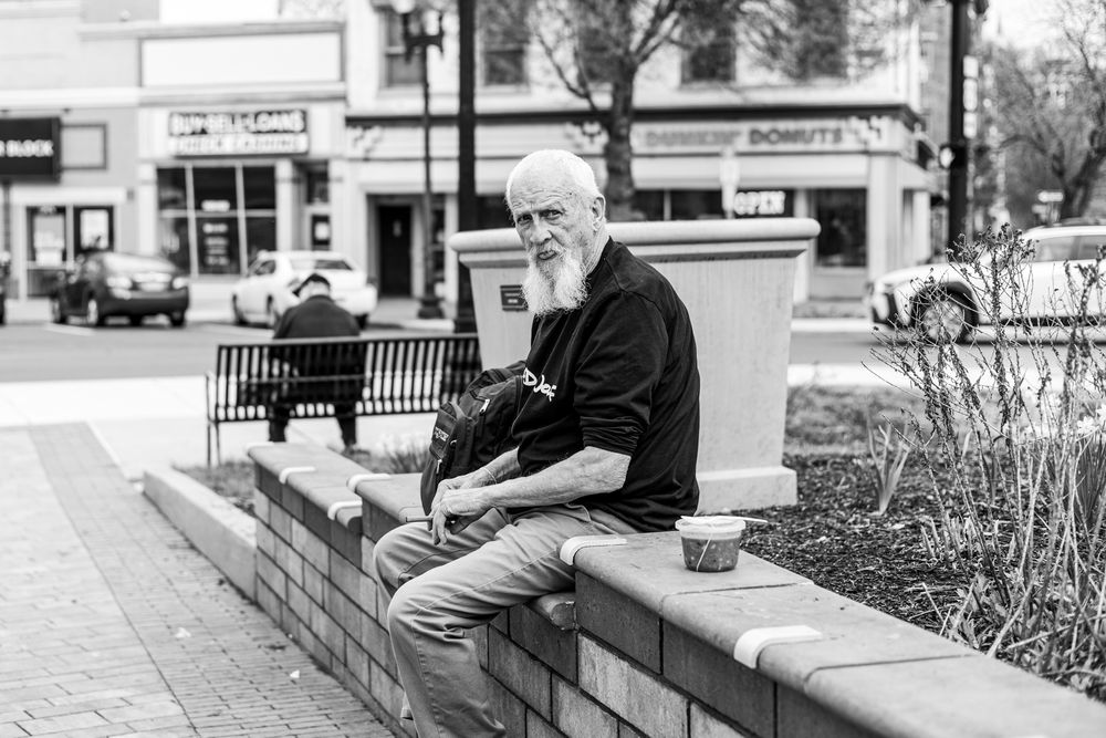 Elderly man with white beard sitting on brick wall in black and white urban street scene with donut shop in background.