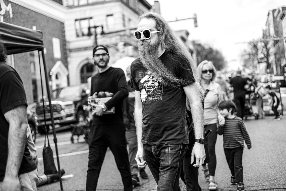 Black and white street photo of long-haired man in sunglasses and graphic tee walking in busy urban intersection.