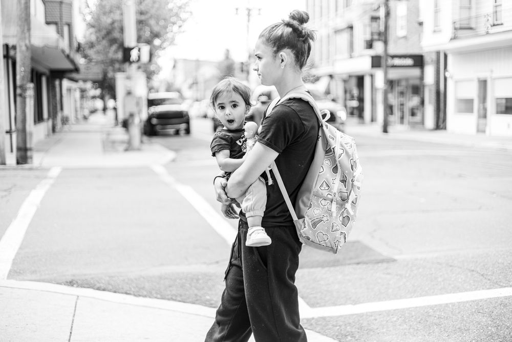 Black and white photo of a young woman carrying a toddler while wearing a backpack, crossing a city street.