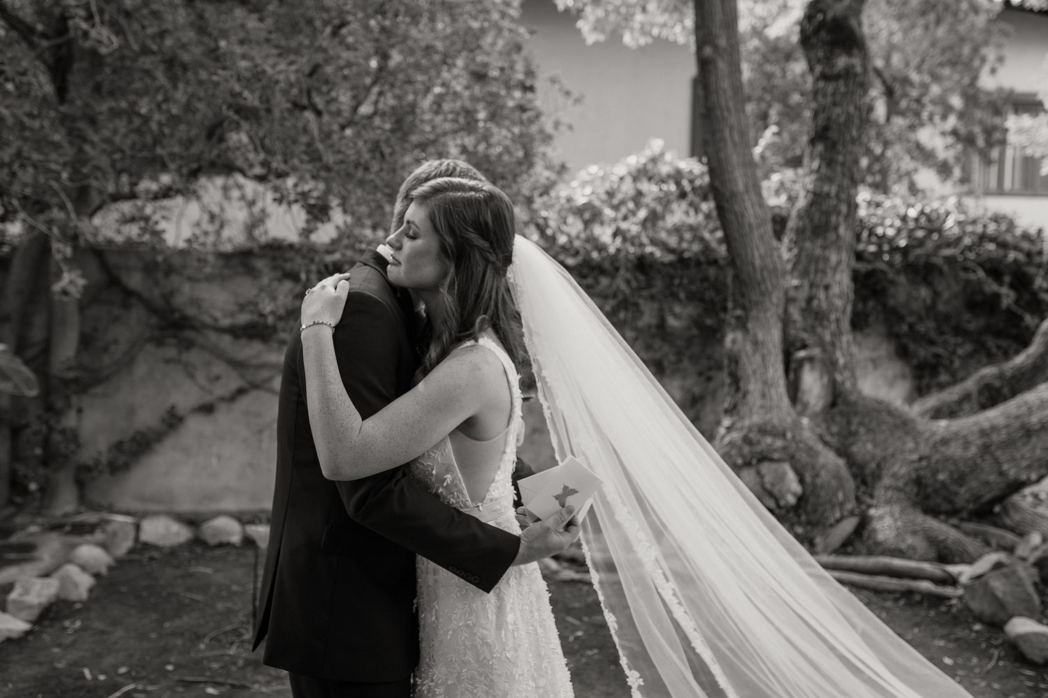Black and white photo of a bride and groom sharing a tender embrace outdoors, with her veil flowing gracefully.