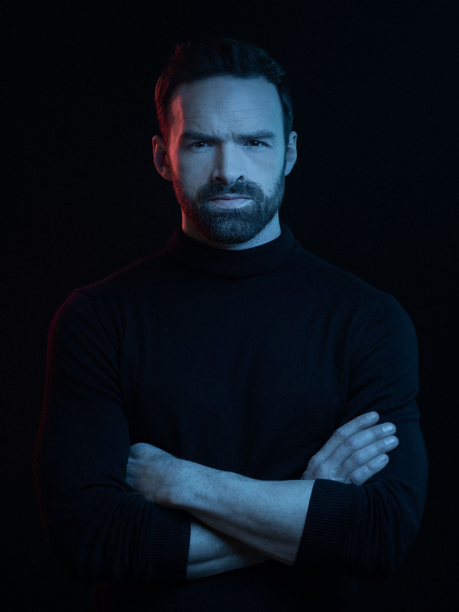 Serious bearded man in black turtleneck with arms crossed, dramatically lit with red and blue lighting on dark background.