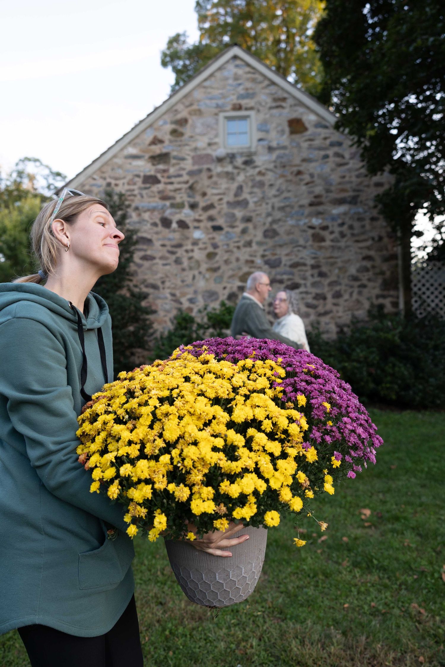 A person carries vibrant yellow and purple mum flowers in pots outside a rustic stone building on an autumn day.