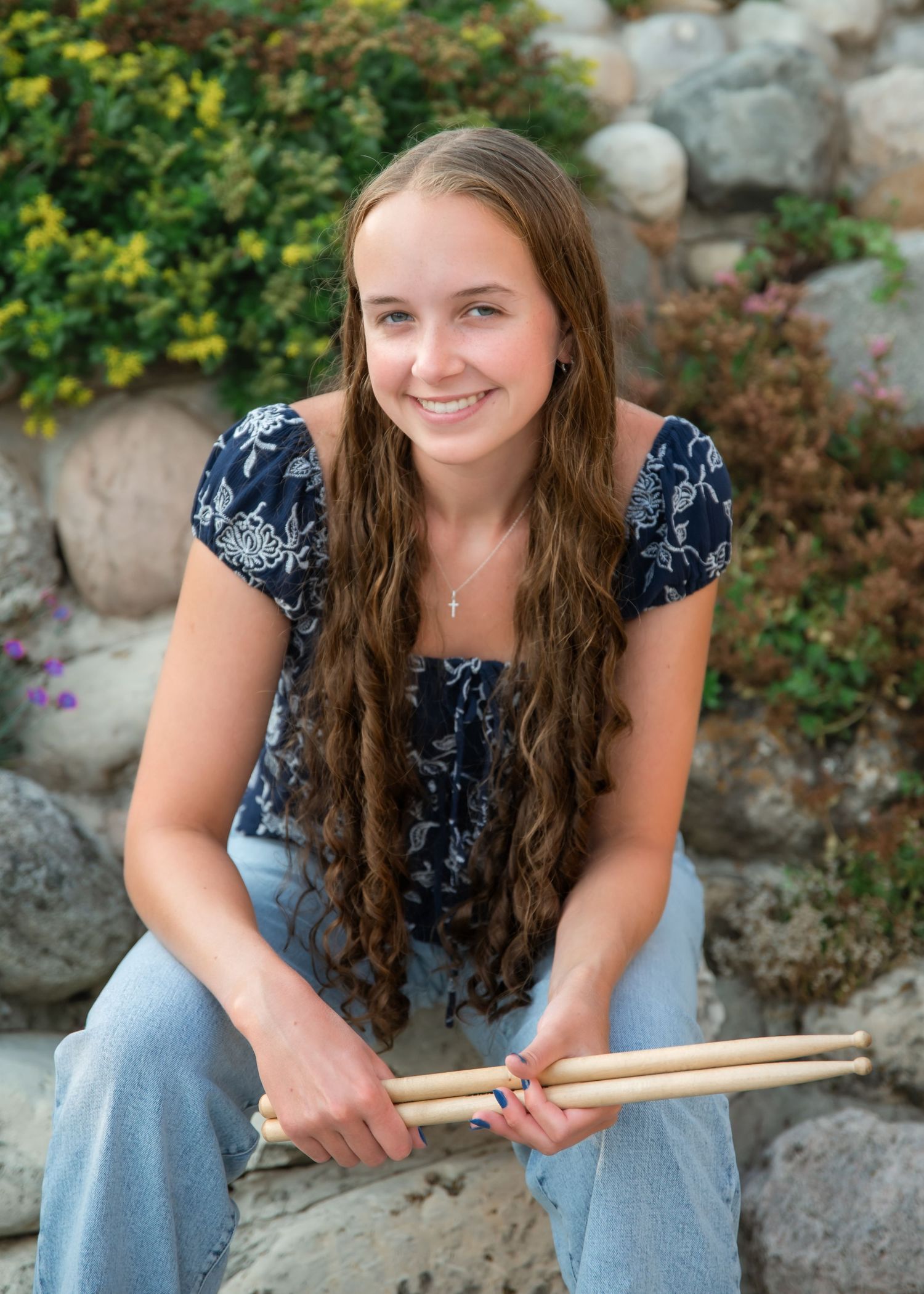 A person in a navy floral top and jeans sits on stone steps while holding drumsticks in an outdoor setting.