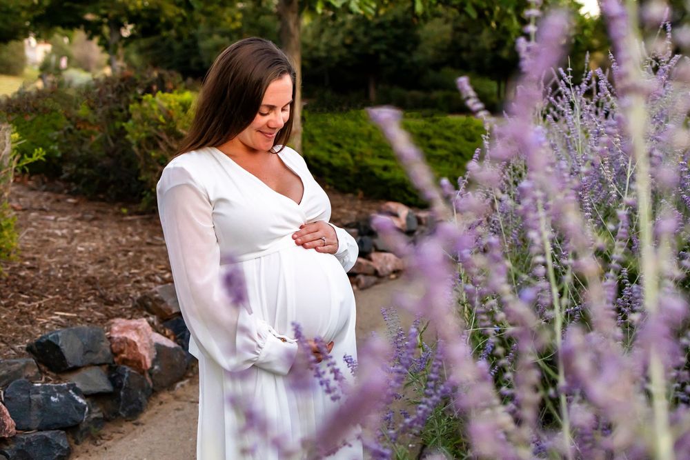 A pregnant woman in a white dress stands among blooming purple lavender flowers in a garden setting.