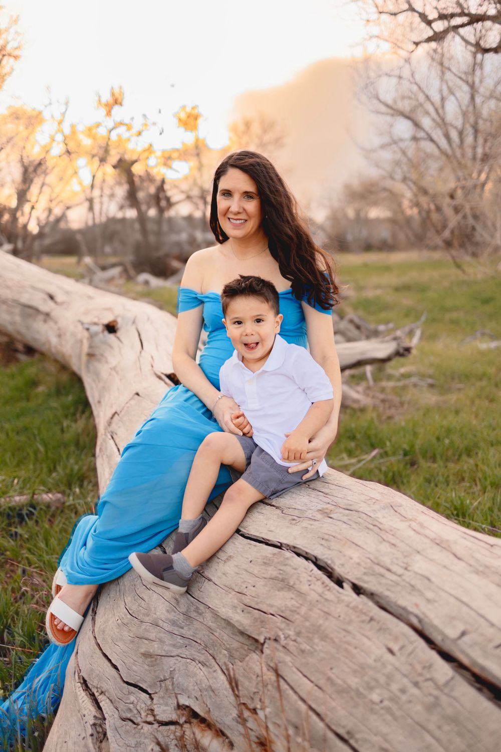 A mother and child share joyful moments together while sitting on a large fallen tree trunk at sunset.