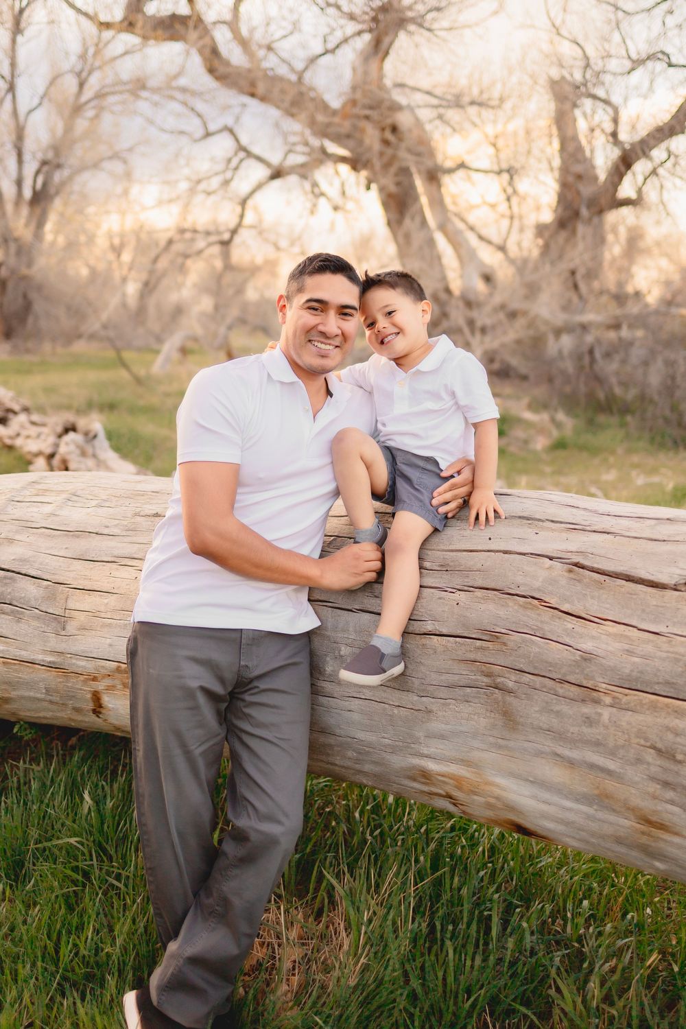 A father and young son share a tender moment while sitting on a fallen log in a natural outdoor setting at sunset.
