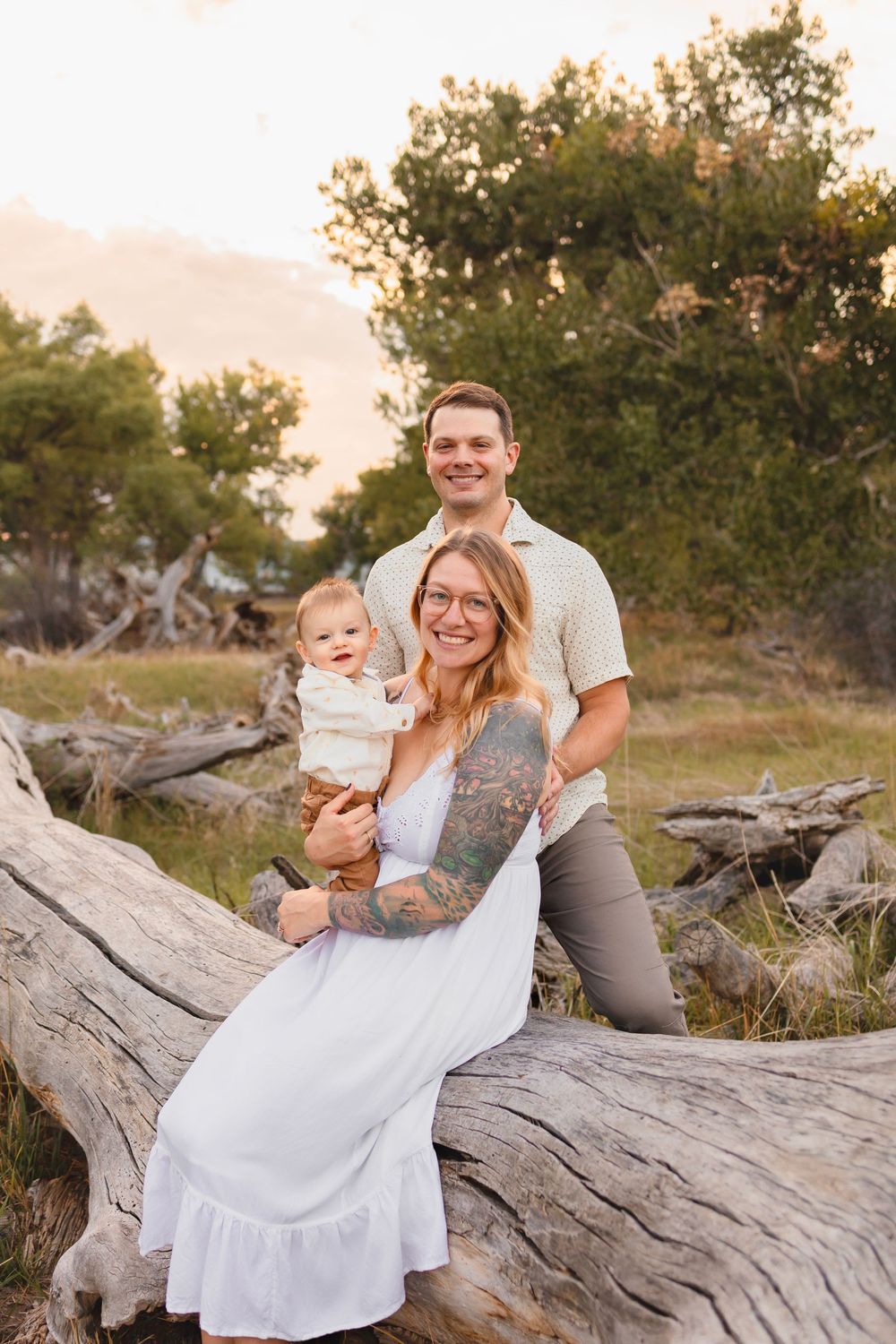 Family enjoying a sunset photo session.  They are sat on a fallen tree in a forest in casual white and gray attire.