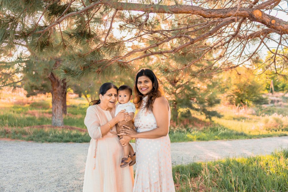 A grandmother holding a baby boy.  He is in the middle between grandmother and his mother.  They are outside underneath a large tree