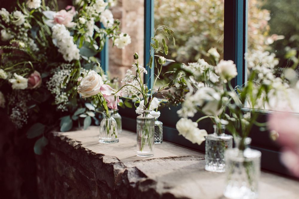 Small glass vases with delicate pink and white flowers and blue candles arranged on a rustic stone windowsill.