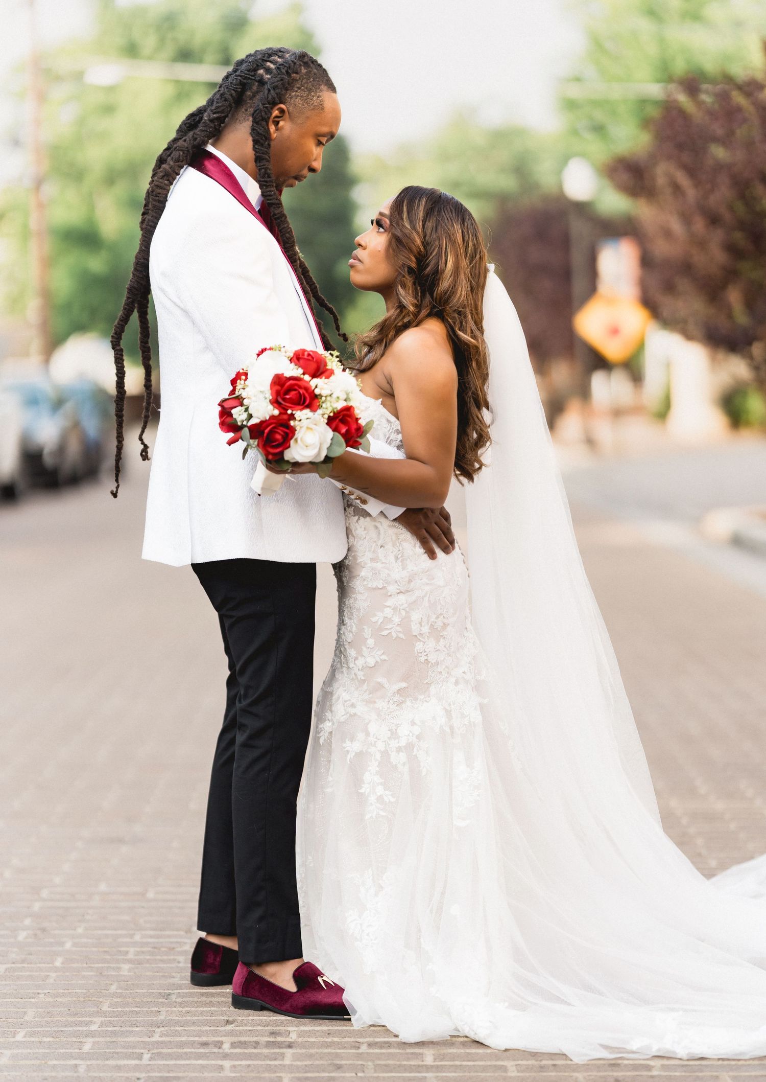 A romantic wedding couple embraces outdoors while wearing a white dress and dark suit with burgundy accents.