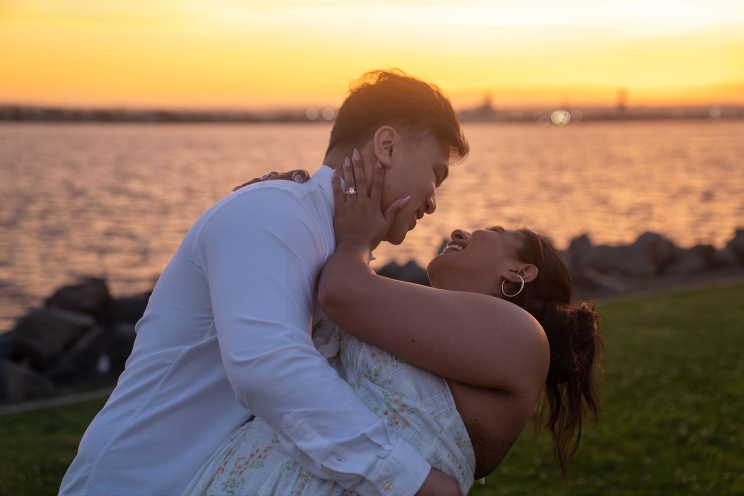 Couple sharing a romantic moment at sunset by the waterfront, embracing and laughing in golden light.