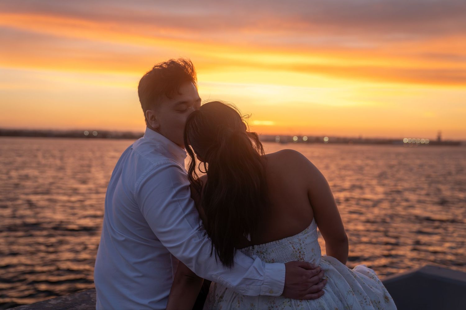 Couple embracing at sunset by the water, sharing a romantic moment with golden orange sky backdrop.