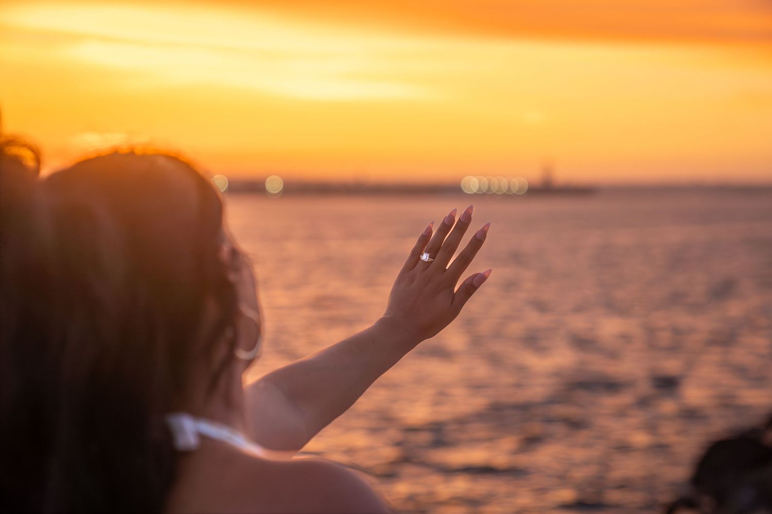 Woman reaching hand toward golden sunset over ocean water with ships visible on horizon.