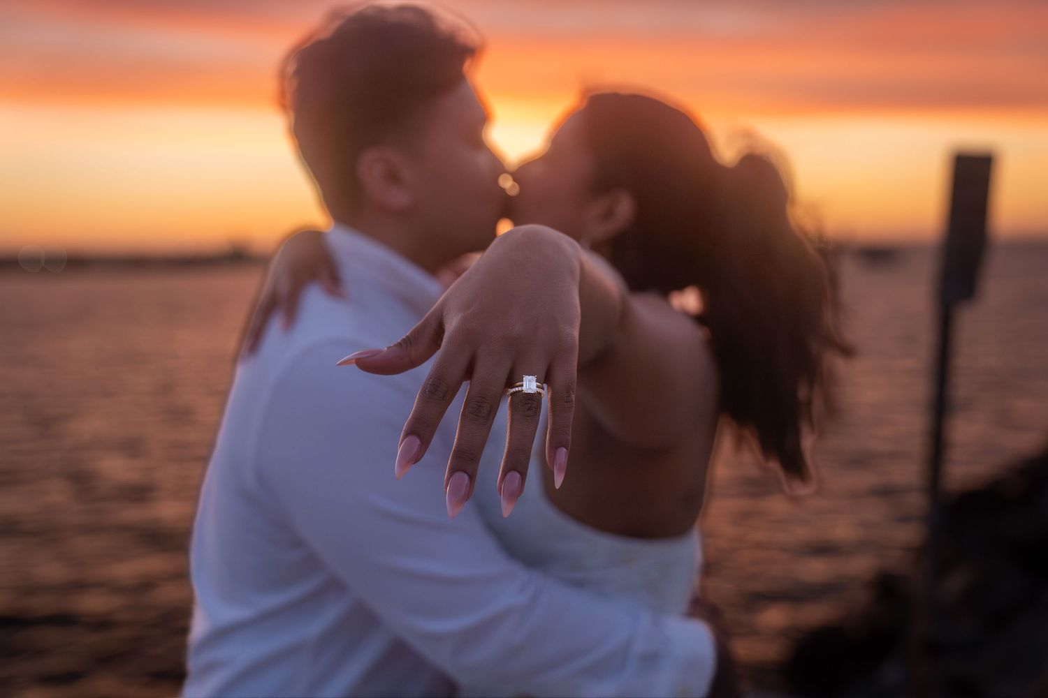 Couple kissing at sunset by the water, woman showing off diamond engagement ring in the foreground.