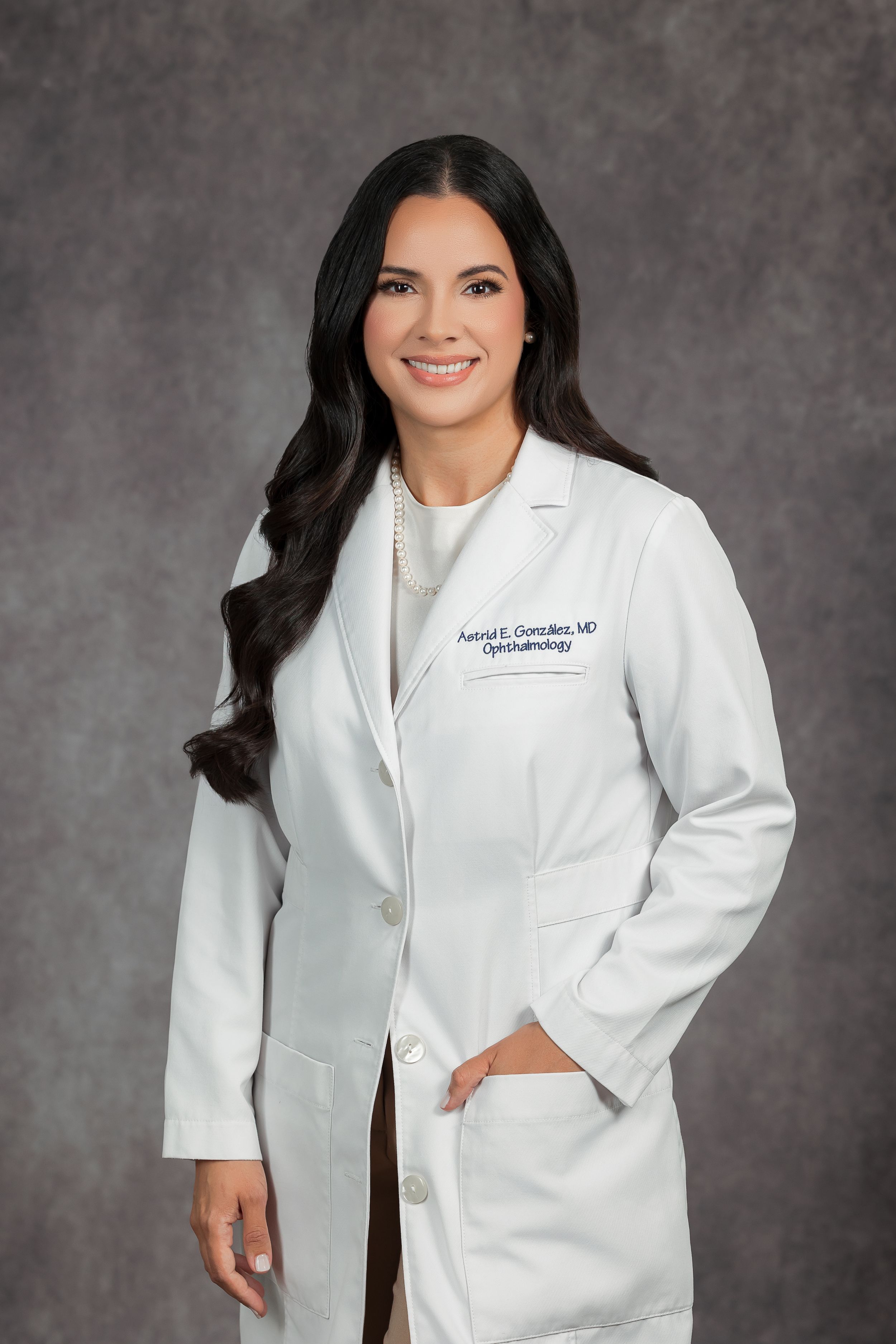A smiling woman wearing a white medical coat stands in front of a gray background.