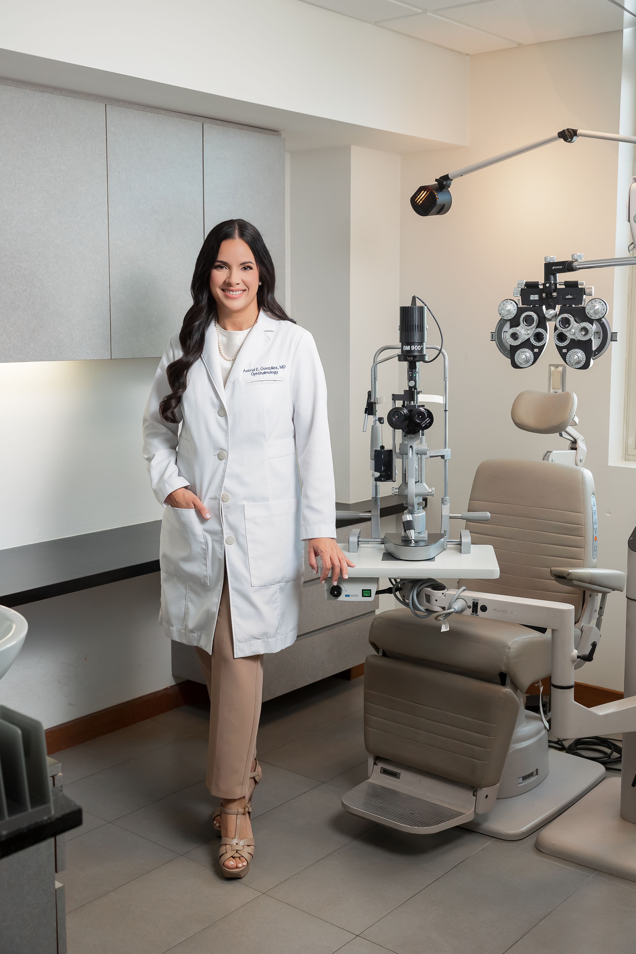 A female optometrist wearing a white lab coat stands in an eye examination room with medical equipment.