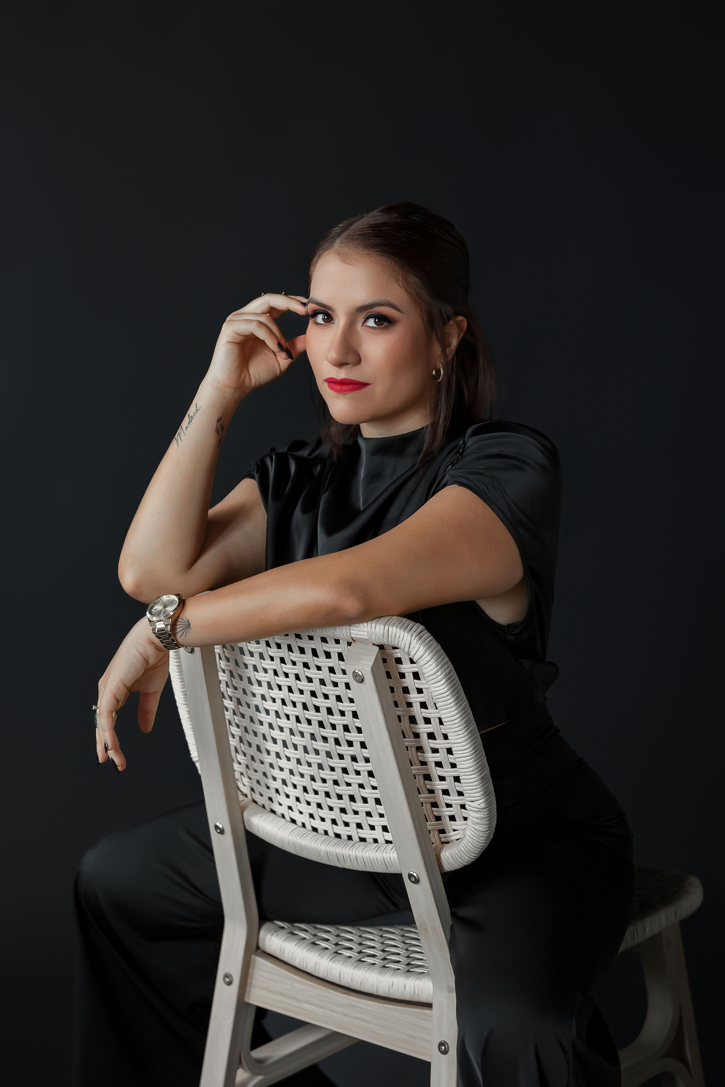 A woman with dark hair and red lips sits on a white chair, looking thoughtfully at the camera against a dark background.
