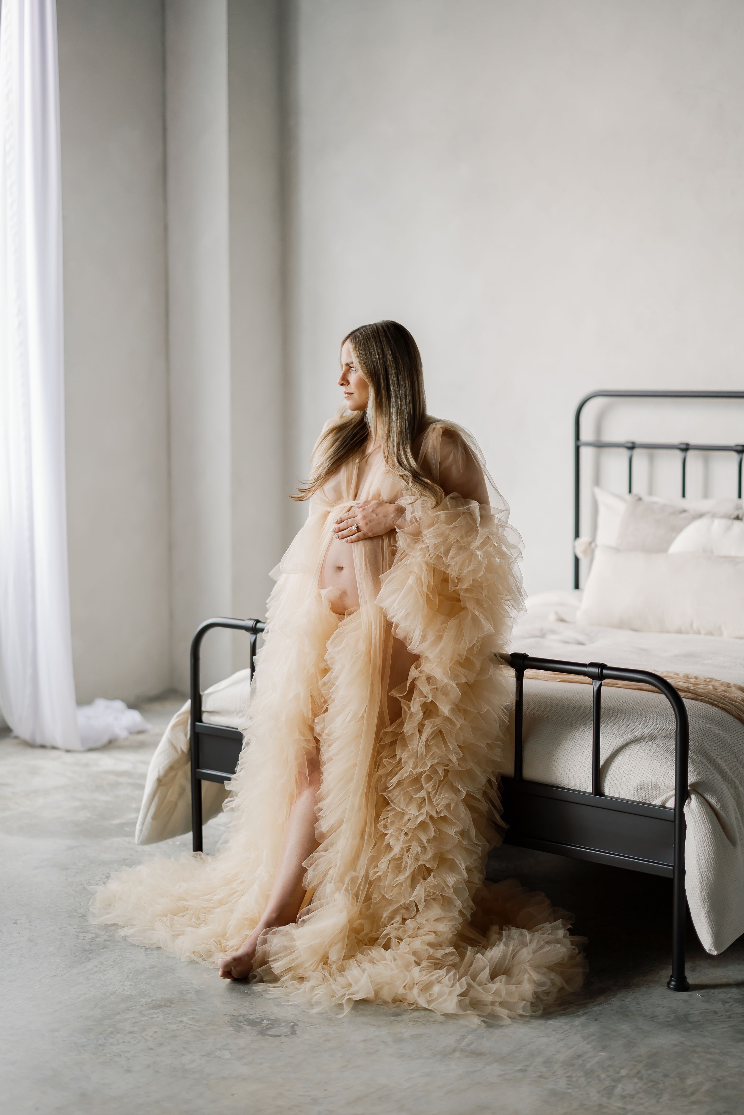 A woman wearing a long, feathery dress sits on a metal bed frame in a minimalist room.