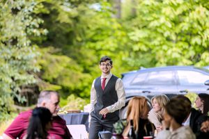 Smiling groomsman in gray vest and red tie walks toward guests seated outdoors at a wedding reception.