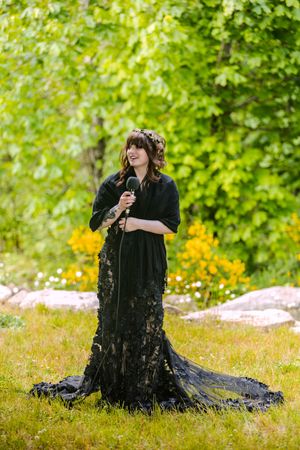 Woman in elegant black lace gown with floral crown holds microphone outdoors in green garden.