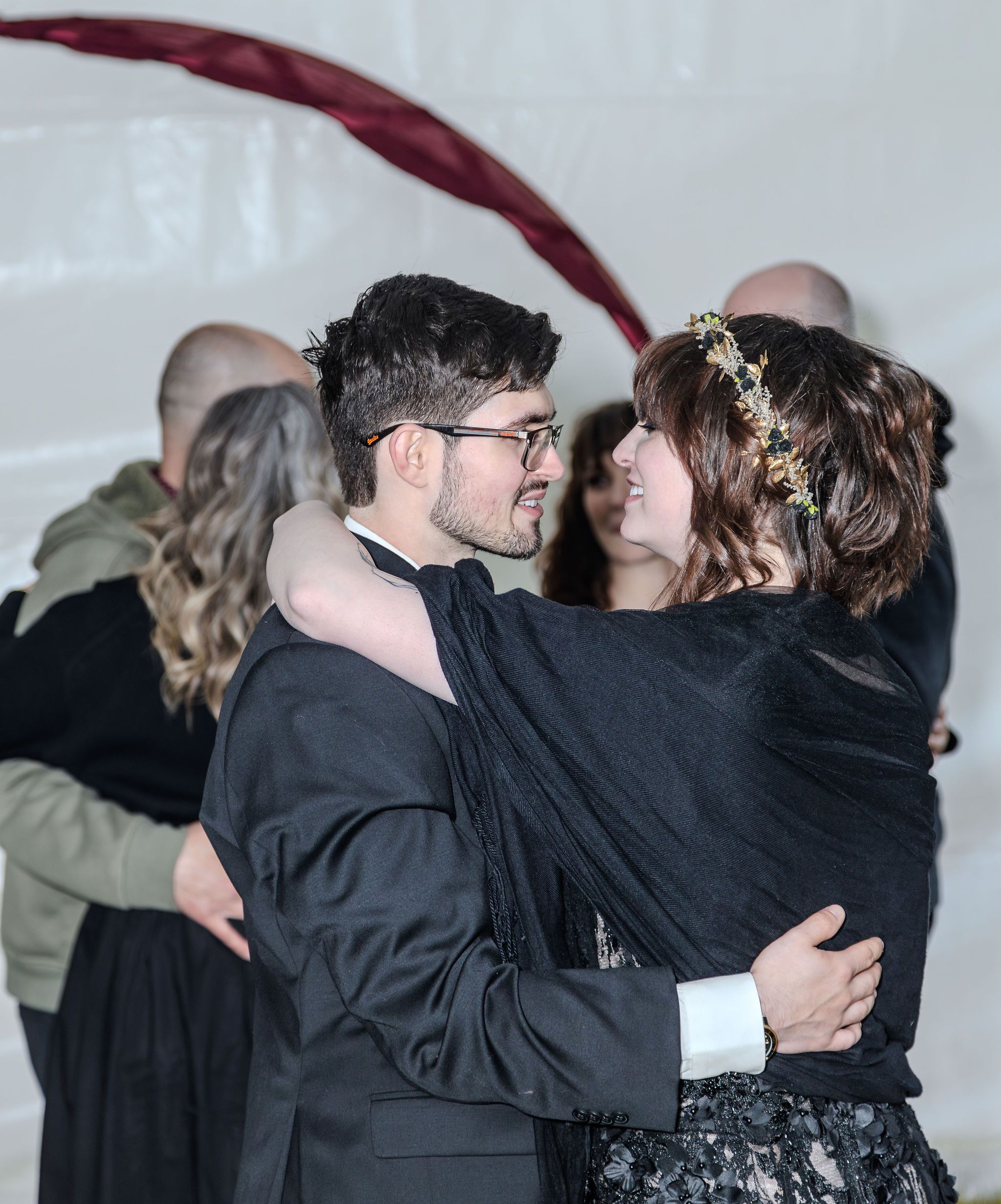 Newlyweds share first dance embrace at wedding reception with guests in background.