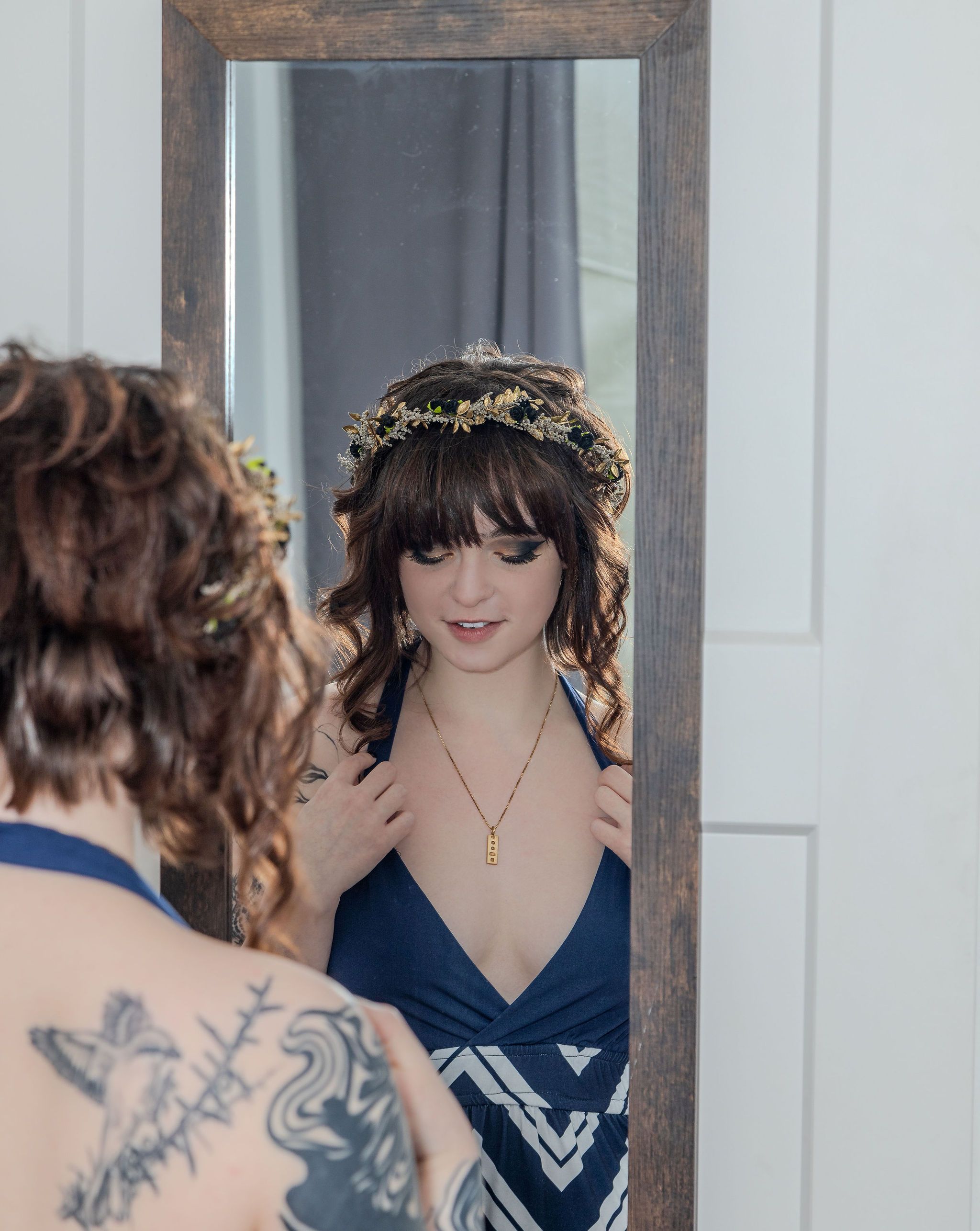 Brunette woman with bangs wearing a floral crown and navy dress admiring herself in a rustic mirror.