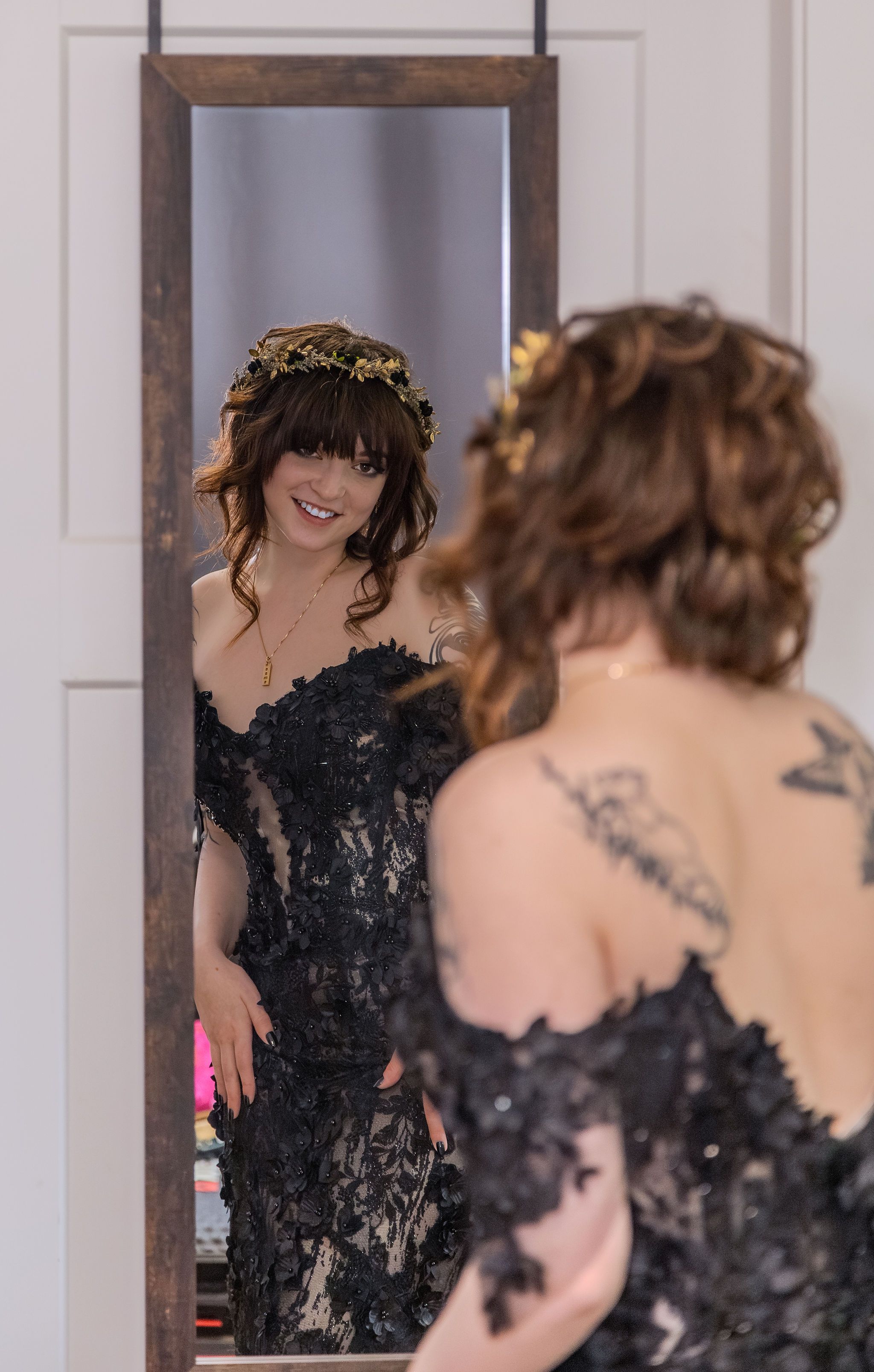 Bride in black lace wedding gown with floral crown admires her reflection in a full-length mirror.