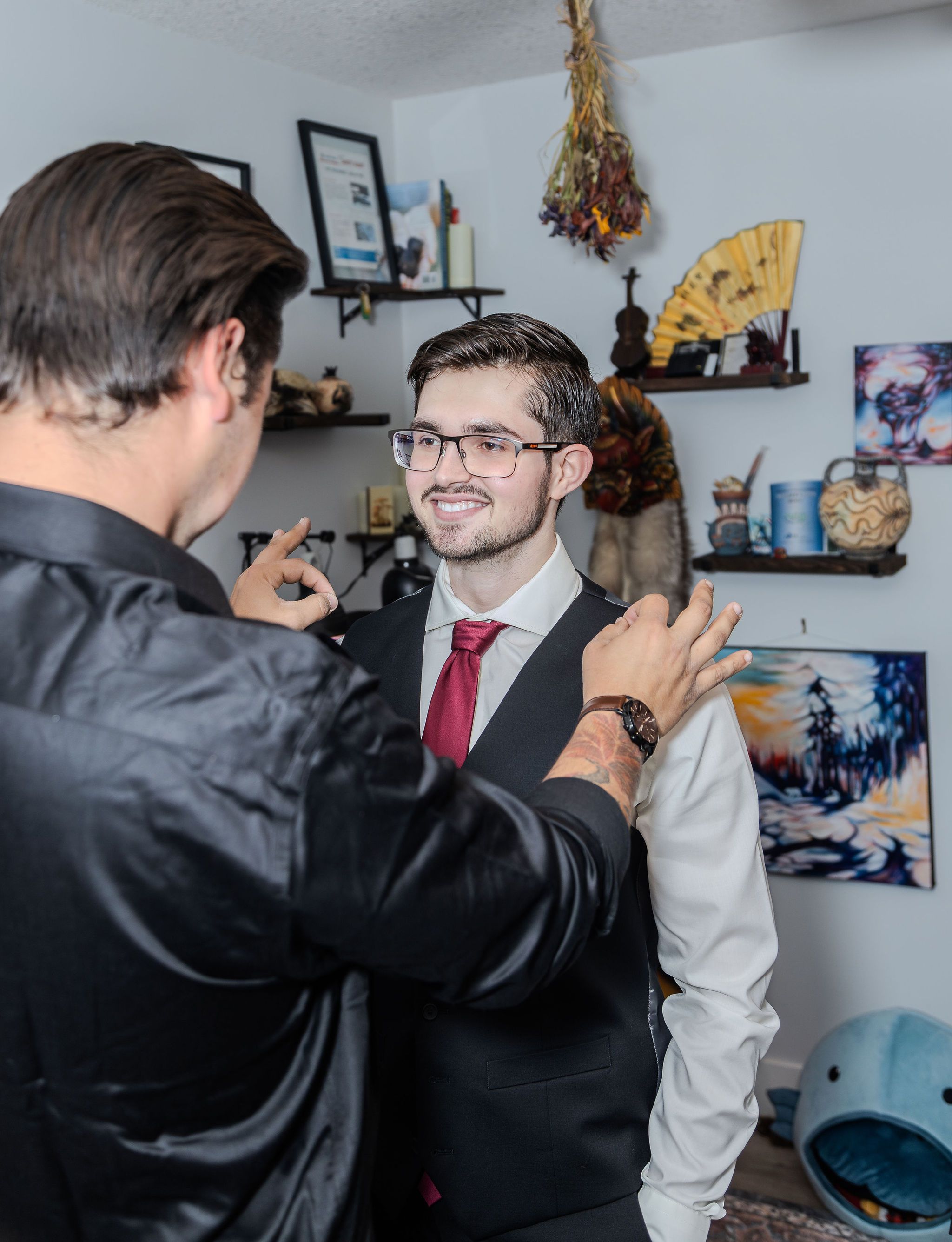 Groom in gray vest and red tie smiles as a friend adjusts his outfit before the wedding ceremony.