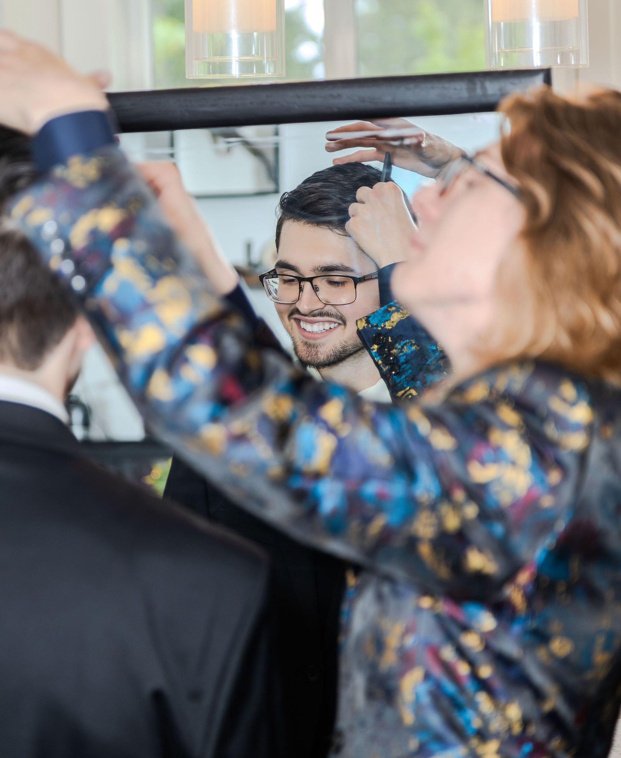 Woman in colorful jacket styles groom's hair while he smiles at his reflection in a mirror.