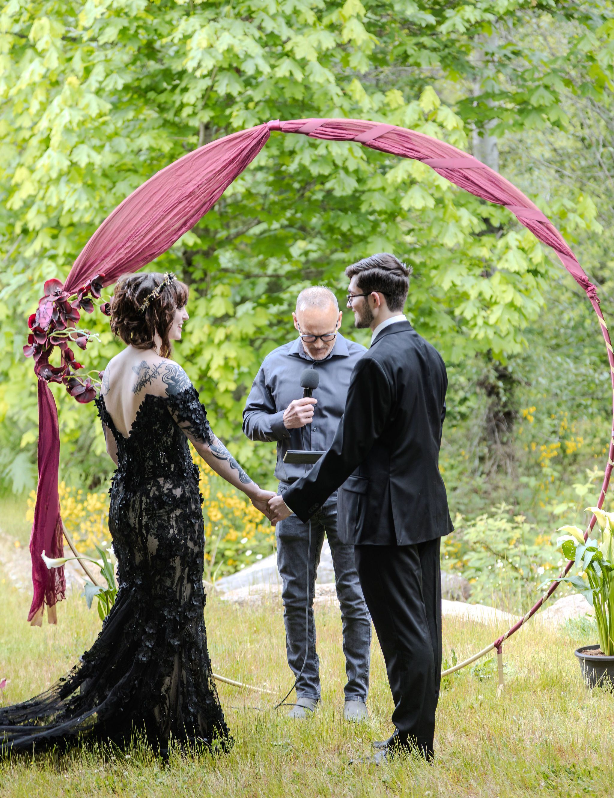 Couple exchanges vows outdoors under a red arch with an officiant in a lush green setting.