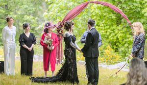 Wedding ceremony outdoors with bride in black gown, groom, officiant, and guests under a red arch.