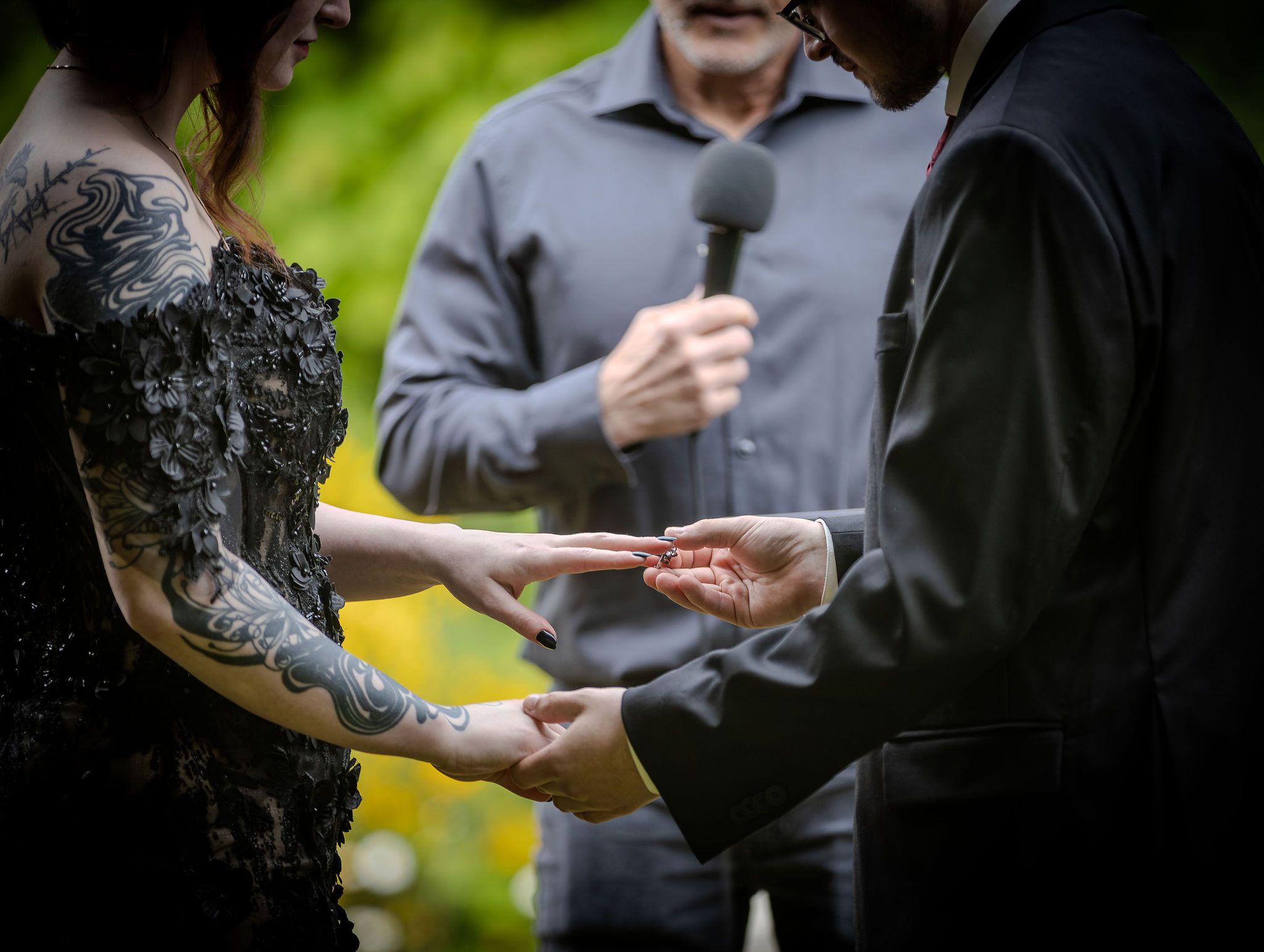 Groom places wedding ring on bride's tattooed hand during outdoor ceremony with officiant behind them.