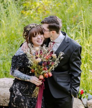 Groom kisses bride's cheek as she smiles holding a wildflower bouquet in a natural outdoor setting.