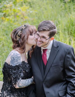 Bride in black lace gown kisses groom's cheek as he smiles in a lush green outdoor setting.