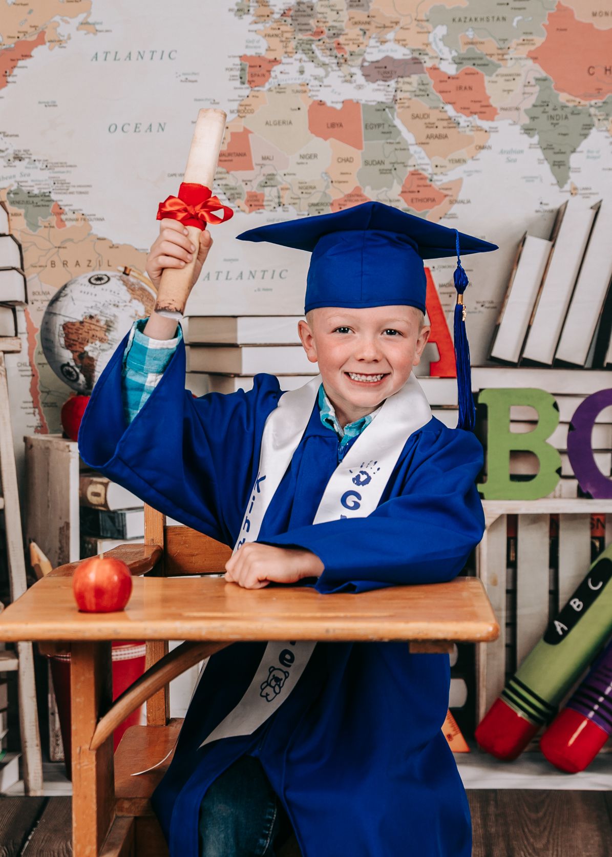 An excited boy in a blue graduation cap and gown holds up a diploma at a desk with a world map backdrop.