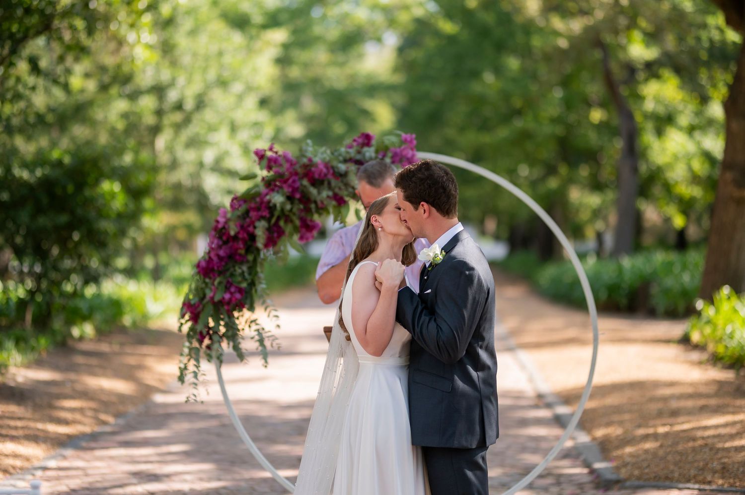 Bride and groom share a kiss under a circular floral arch adorned with purple flowers on a tree-lined path.