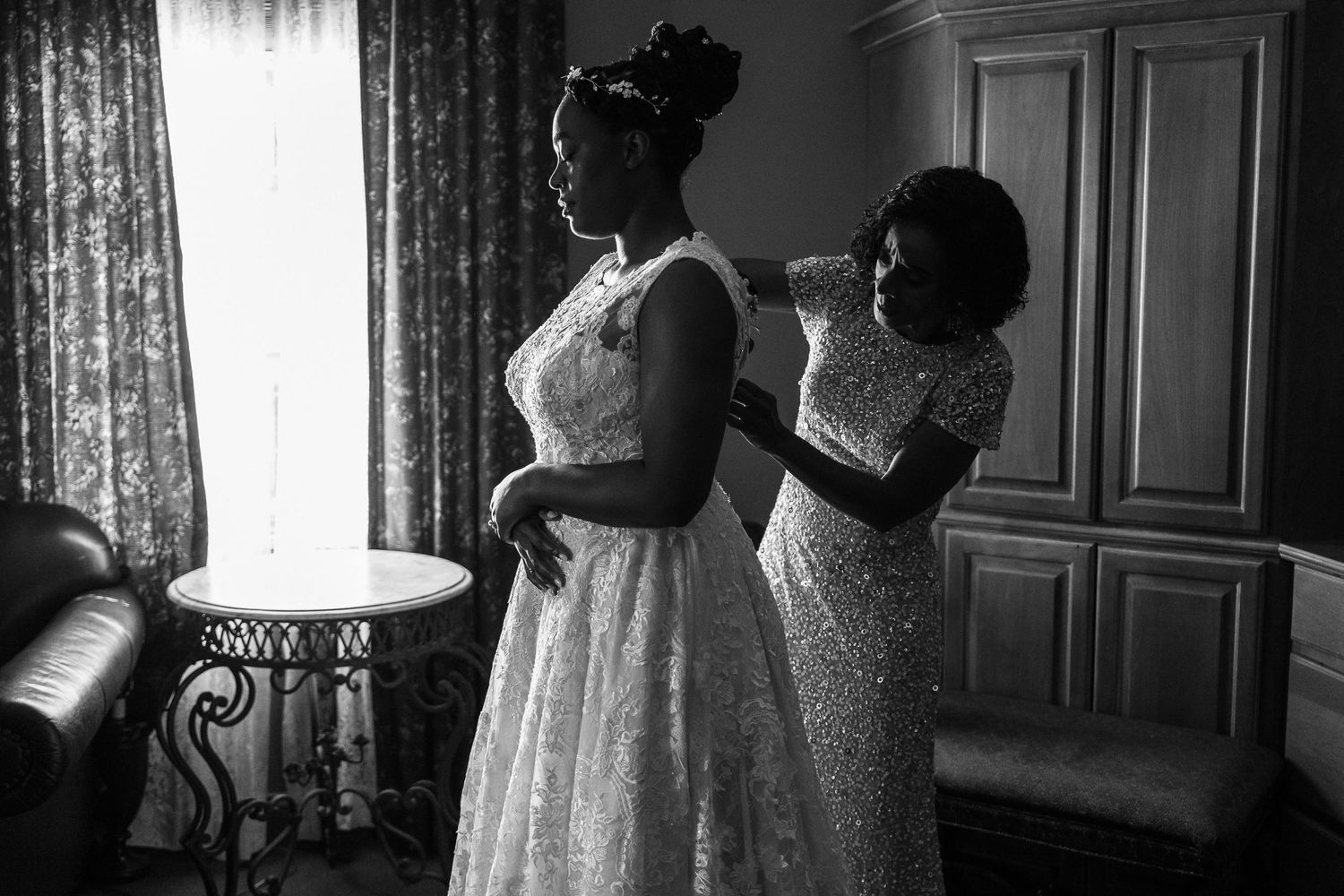 Black and white photograph showing figures in elegant dresses standing by curtained windows in a vintage interior setting.