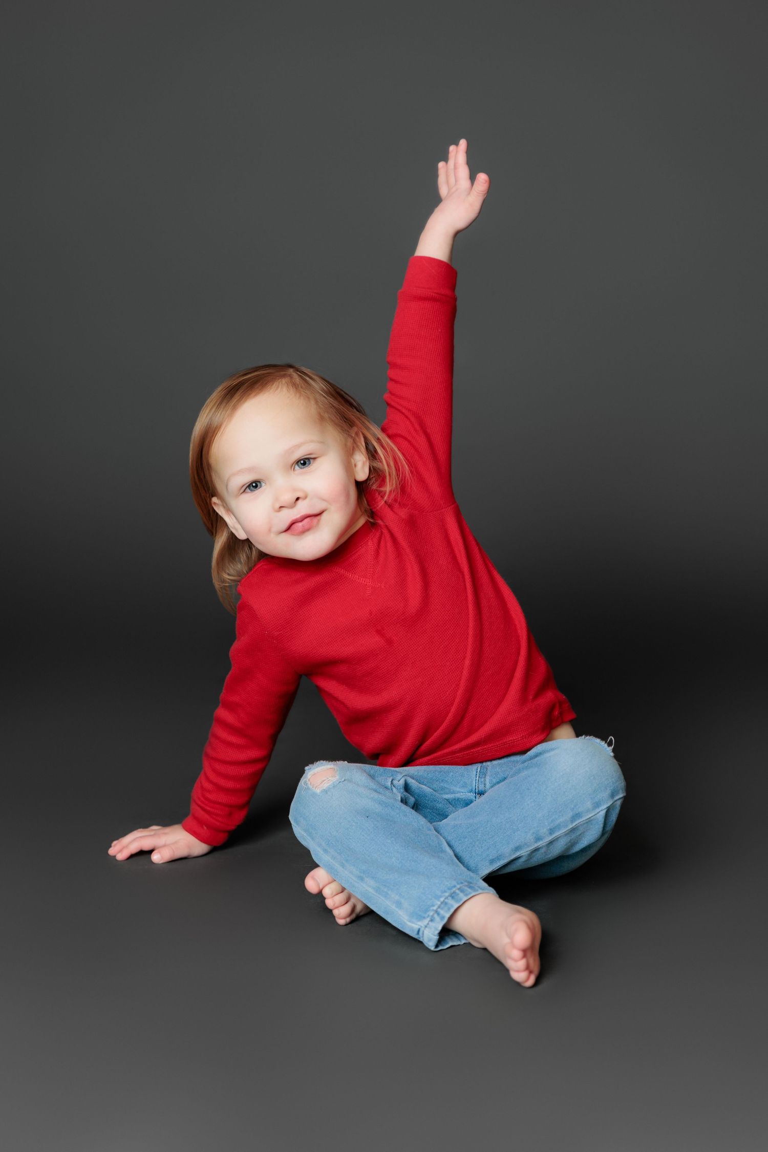 A child in a red sweater and jeans plays and poses playfully against a dark gray studio backdrop.
