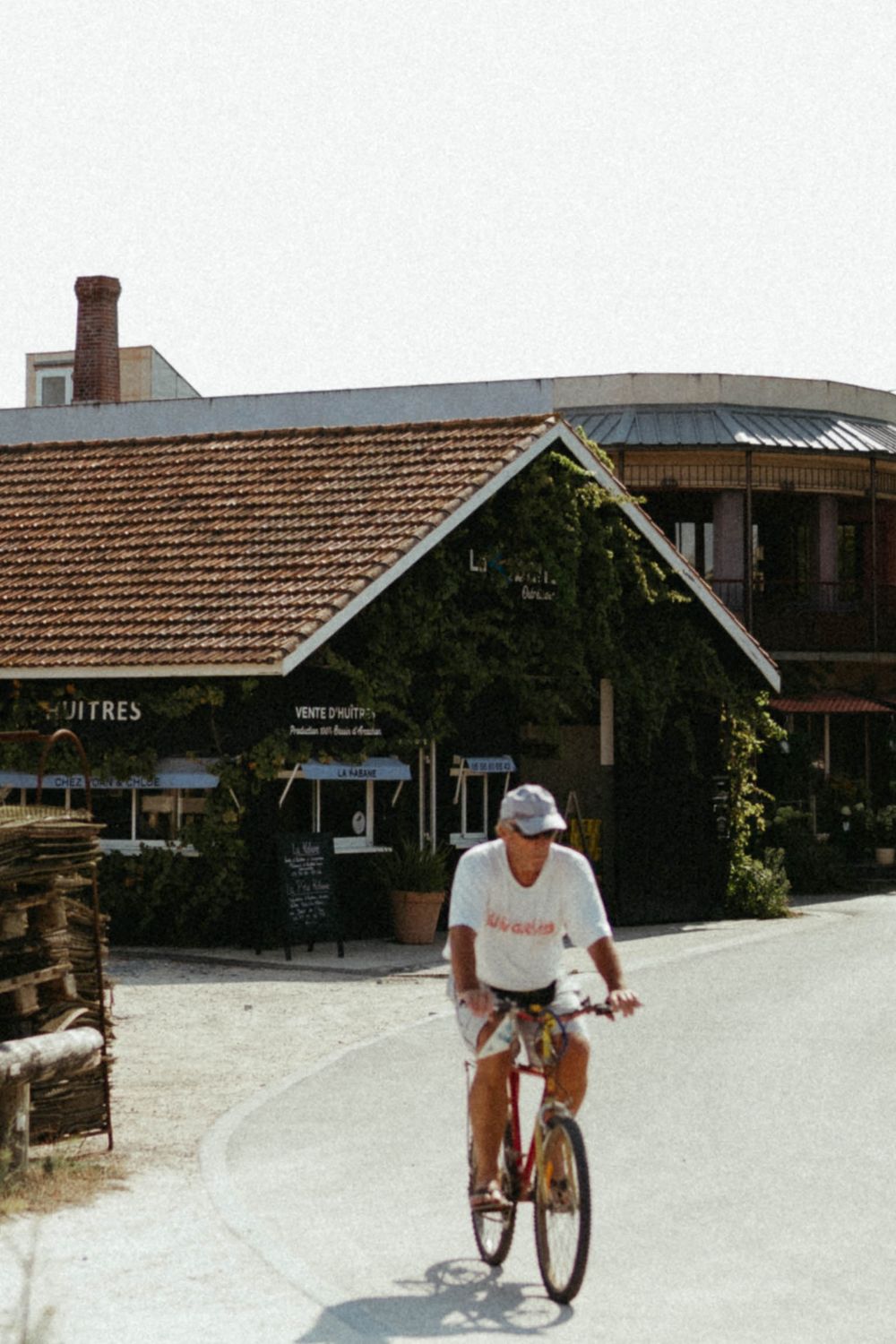 Man in white t-shirt and cap cycling past a French oyster shop on a sunny day.