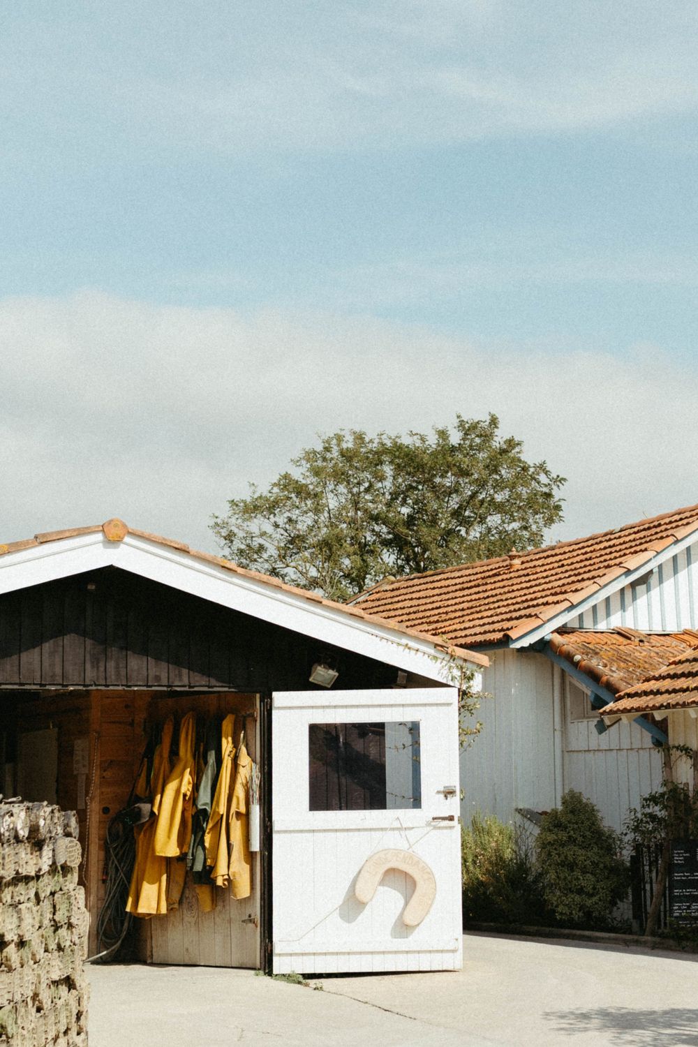 Rural farm buildings with yellow raincoats hanging outside, a white shed with window, terracotta roof tiles, and a tree under blue sky.