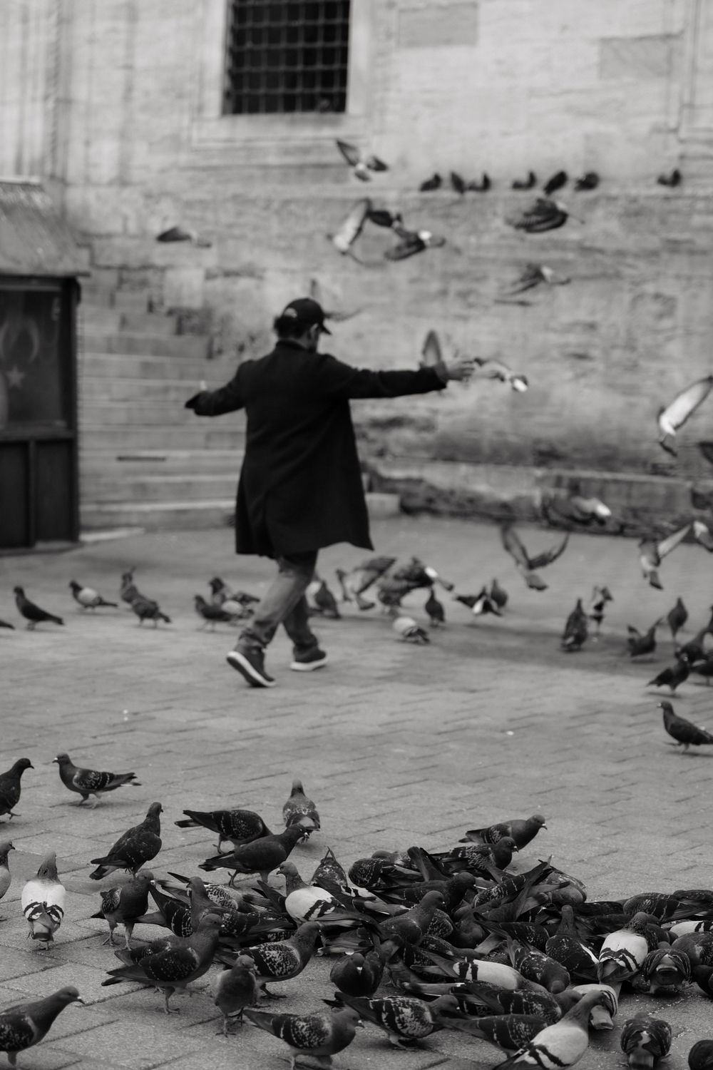 Black and white photo of a person in a coat feeding pigeons in a city square, with birds flying around them.