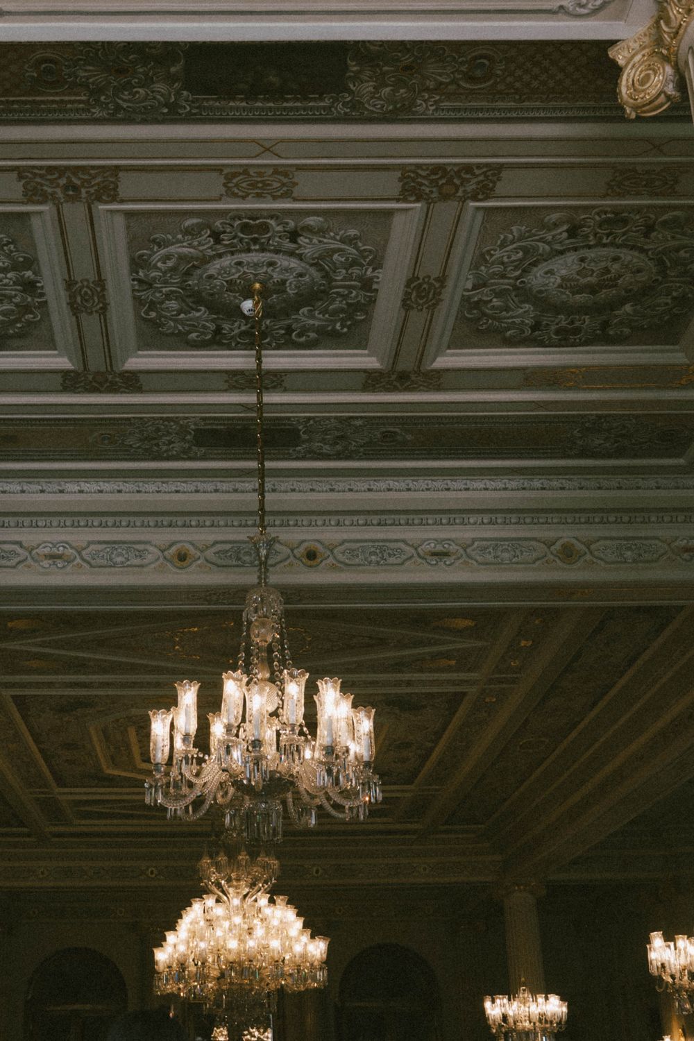 Ornate coffered ceiling with intricate moldings and two elegant crystal chandeliers illuminating a grand ballroom.