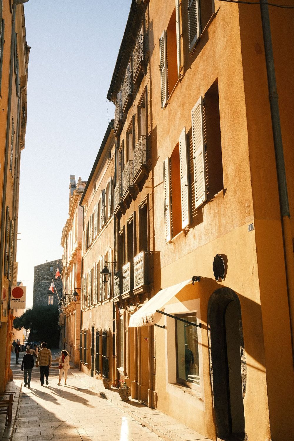 Sunlit narrow street in Aix-en-Provence with warm ochre buildings, wooden shutters, and pedestrians strolling on cobblestones.