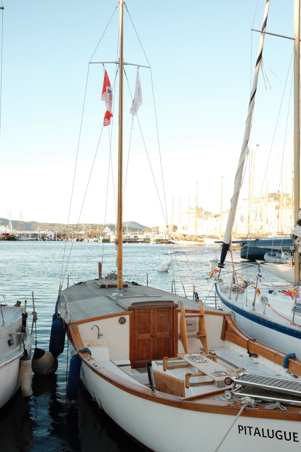 Sailboat named Pitalugue docked at a sunny marina with flags flying from its mast and wooden cabin details.