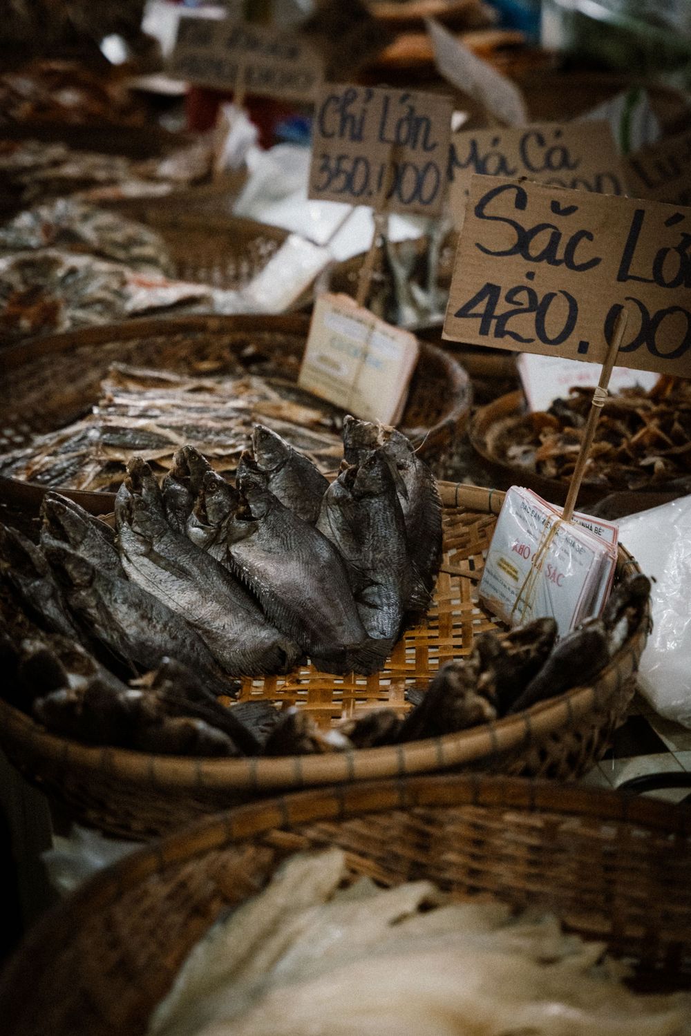 Dried fish displayed in woven baskets at a Vietnamese market with handwritten price signs showing 420,000 VND.