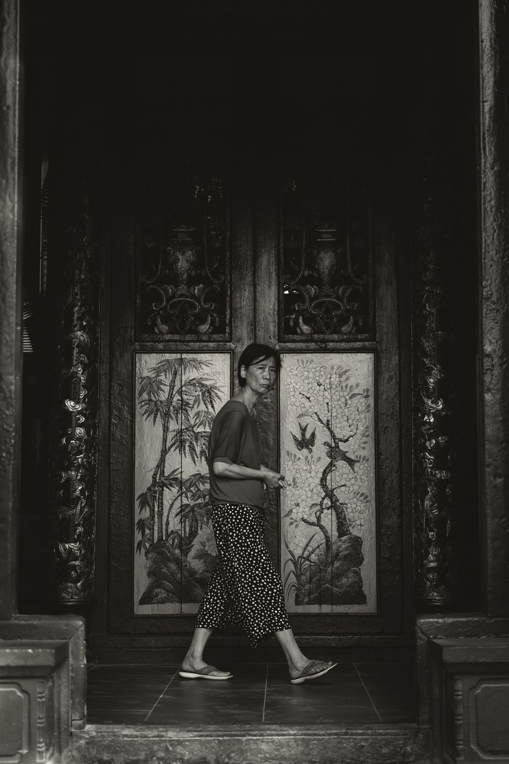 Black and white photo of a woman walking past ornate carved wooden panels with nature motifs in a traditional Asian building.