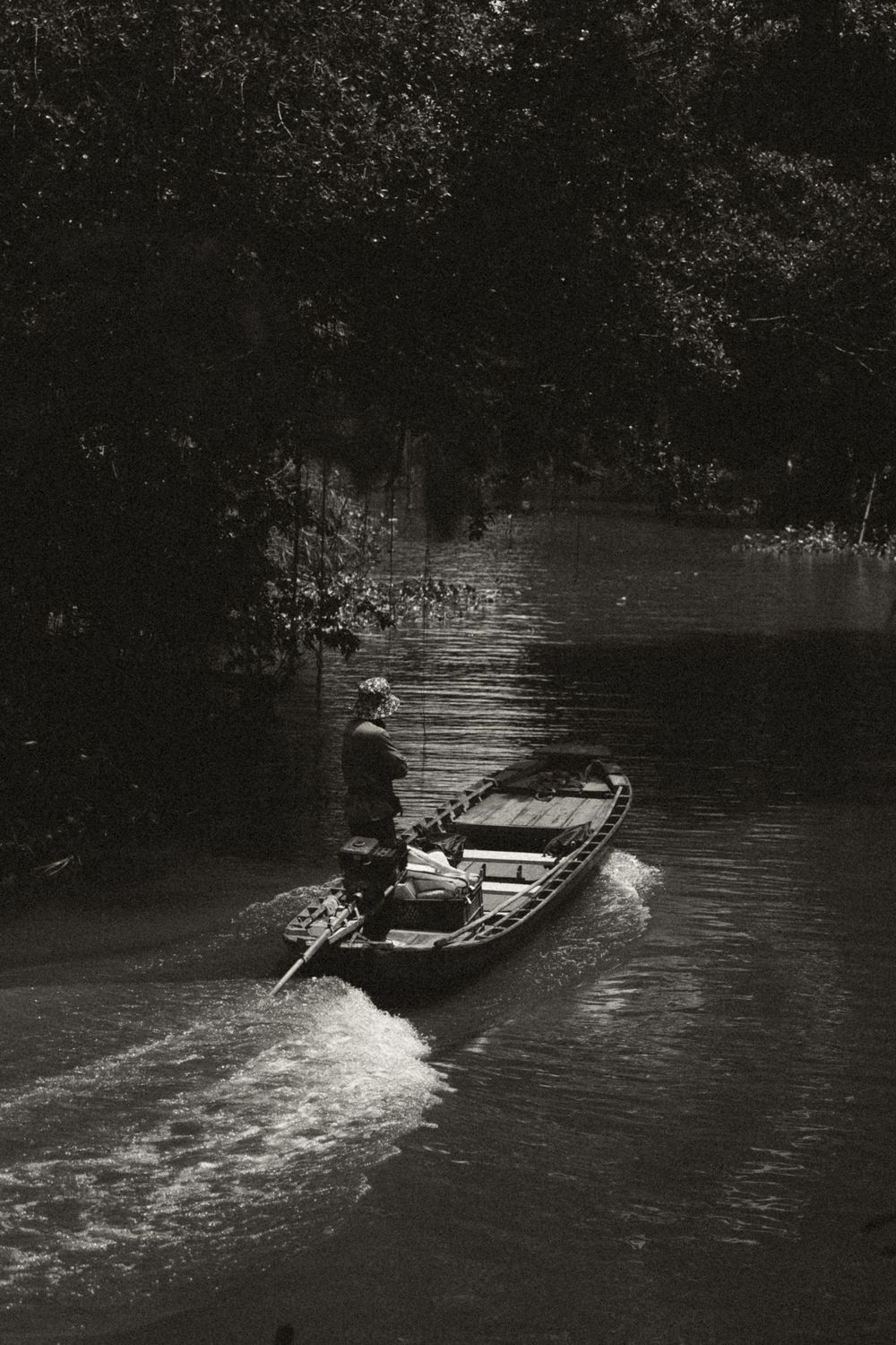 Photo en noir et blanc d'une personne dirigeant un bateau à moteur en bois à travers un canal fluvial étroit bordé d'arbres.